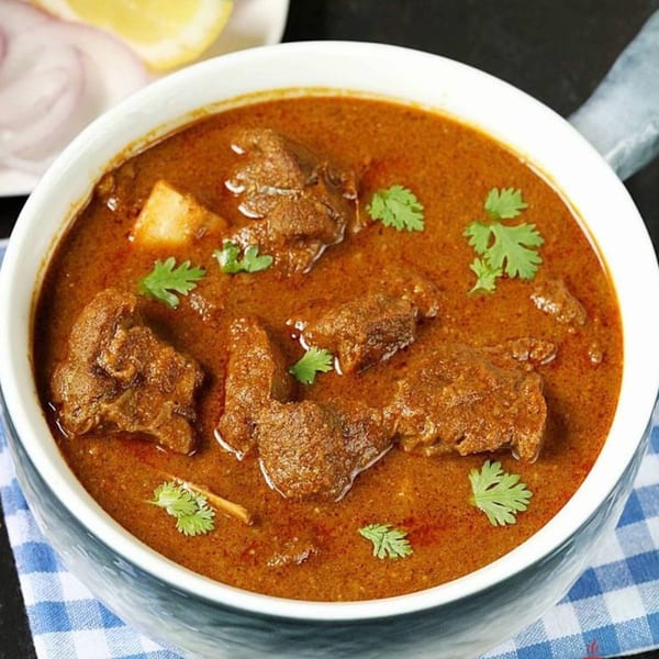 A bowl of rich, brown curry with chunks of meat and fresh cilantro, served with onions and lemon.