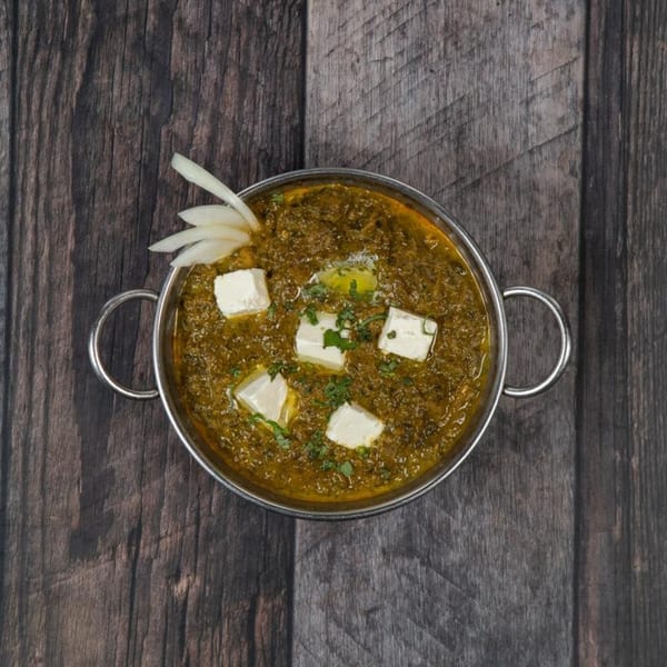 Overhead view of Saag Paneer with paneer cubes, butter, and onion garnish in a metal bowl.