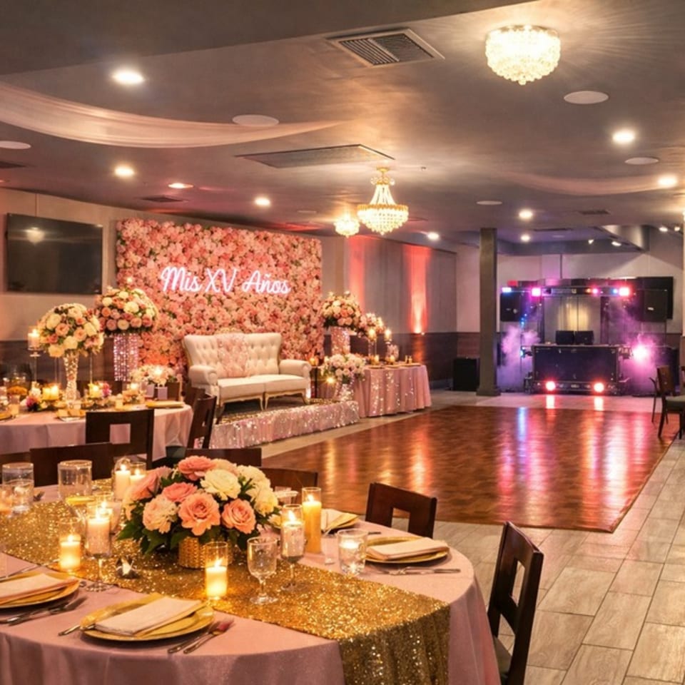 Elegant event hall with pink tables, floral wall with 'Mis XV Años' sign, dance floor, and DJ setup.