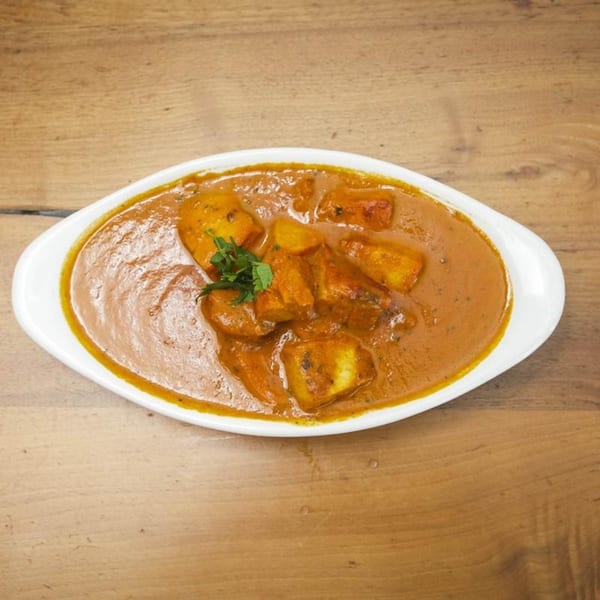 A white oval dish filled with a rich orange curry, paneer, and potatoes, garnished with herbs.