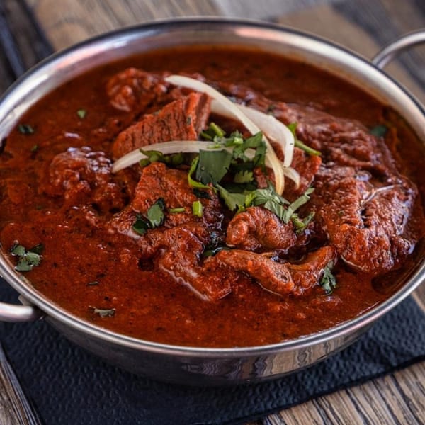 Rich red curry with meat, garnished with fresh cilantro and sliced onions in a metal bowl.