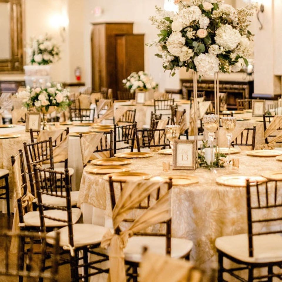 Elegant event space with multiple round tables draped in gold, set with charger plates and floral centerpieces.